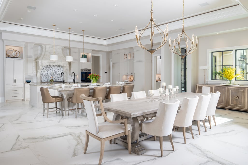 Luxury open-concept kitchen and dining room by Romanza Interior Design featuring white cabinetry, marble flooring, statement chandeliers, and neutral furnishings.